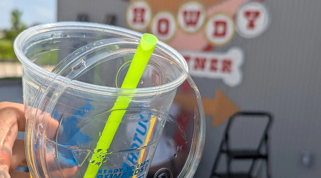 A cup is held in the hand by a person standing in front of a mural painting in Houston, Texas. The green straw is the most vibrant color against the grey wall of the mural.